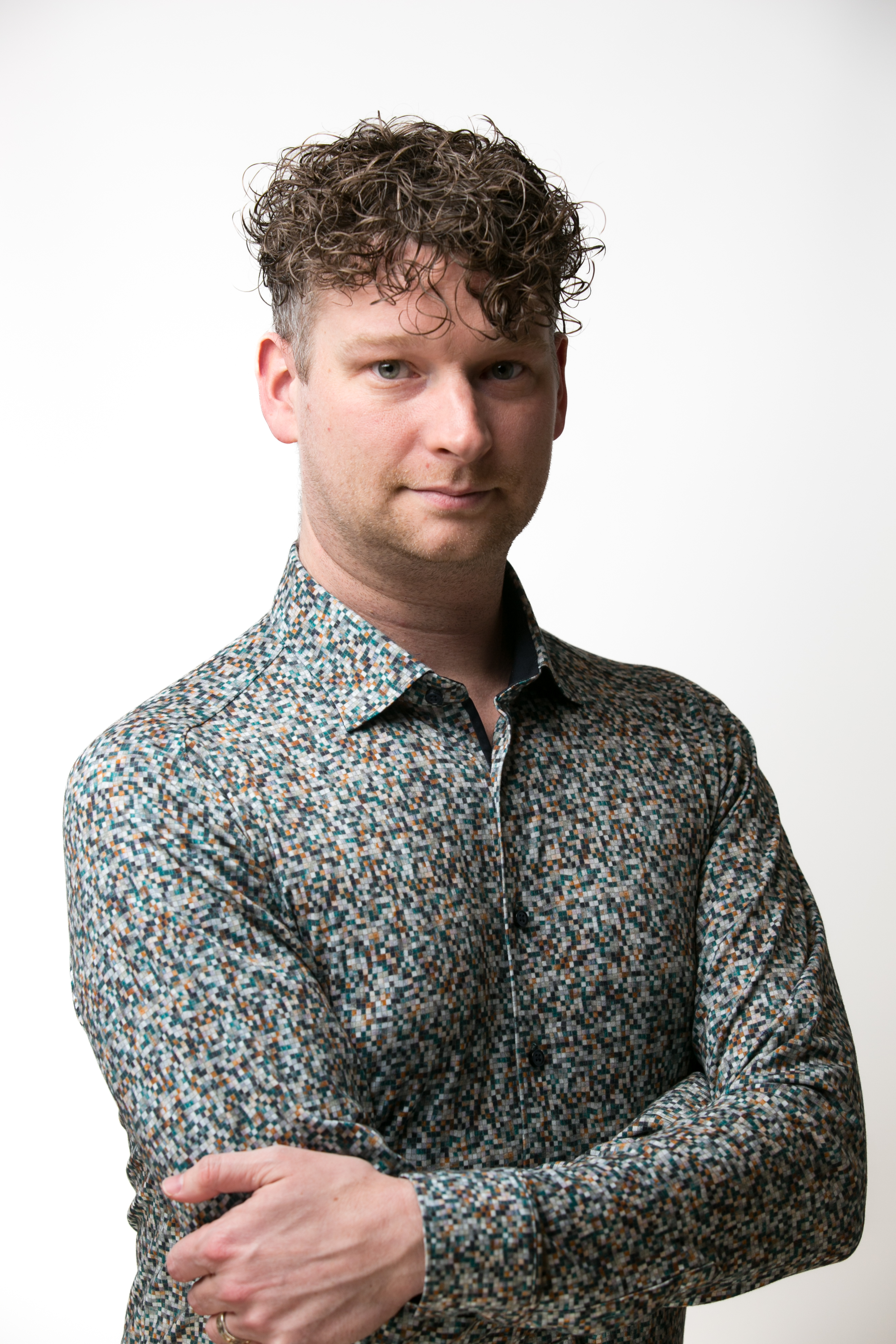 A man with curly brown hair and a multicolored shirt looks at the camera with a neutral expression. His right hand is tuck...